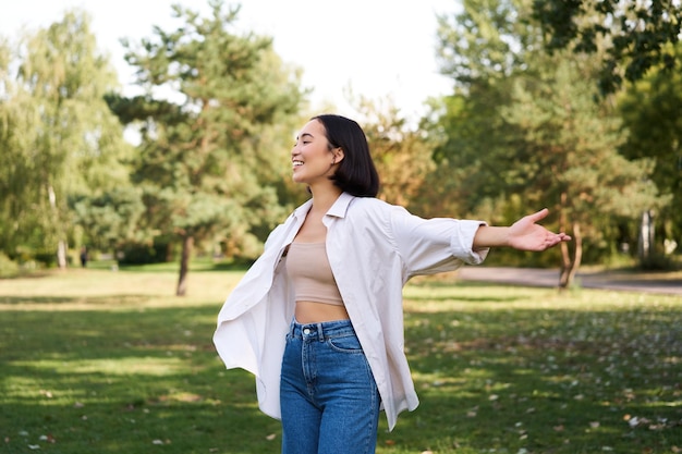 Chica asiática despreocupada riendo y bailando en el parque disfrutando de un día soleado de verano levantando las manos y brea