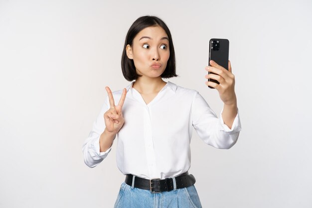 Una chica asiática divertida que muestra un signo de paz en la cámara del teléfono inteligente posando para tomar fotos con una aplicación móvil de pie sobre fondo blanco