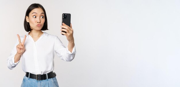 Chica asiática divertida que muestra el signo de la paz en la cámara del teléfono inteligente posando para tomar fotos con mobi