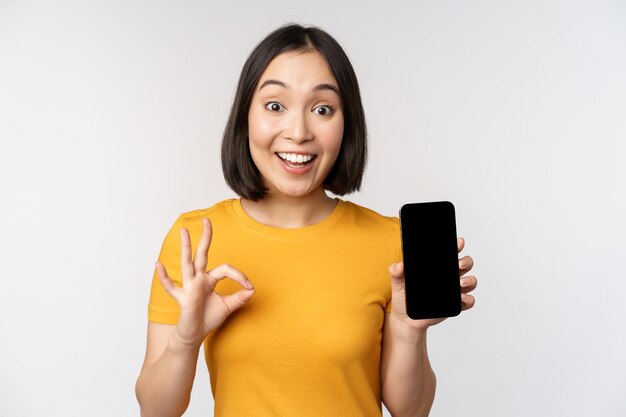 Chica asiática emocionada mostrando la pantalla del teléfono móvil bien señal recomendando la aplicación de teléfono inteligente de pie en camiseta amarilla sobre fondo blanco