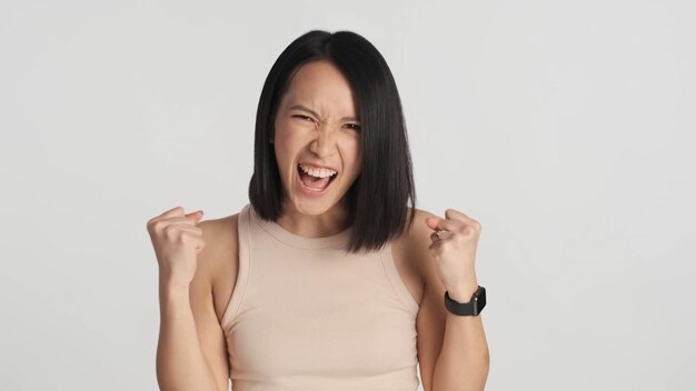 Chica asiática emocional que parece feliz regocijándose de ganar en la cámara sobre el fondo Mujer exitosa