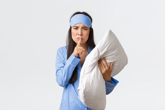 Chica asiática enojada disgustada con máscara para dormir y pijama, sosteniendo la almohada y frunciendo el ceño molesto, haciendo silencio, diciendo shhh cuando se despertó por un ruido fuerte, pidiendo a la gente que se callara por la noche, fondo blanco.