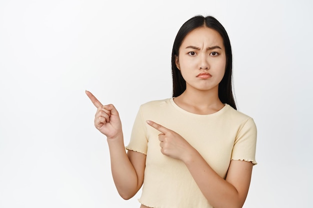 Una chica asiática enojada y malhumorada señalando con el dedo a la izquierda frunciendo el ceño disgustada quejándose de algo de fondo blanco