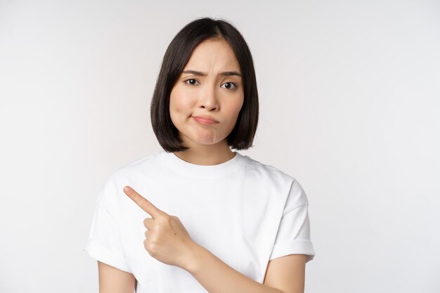 Chica asiática escéptica con camiseta blanca que señala el producto o el logotipo con una mueca decepcionada que no le gusta y se queja de algo de pie sobre fondo blanco.