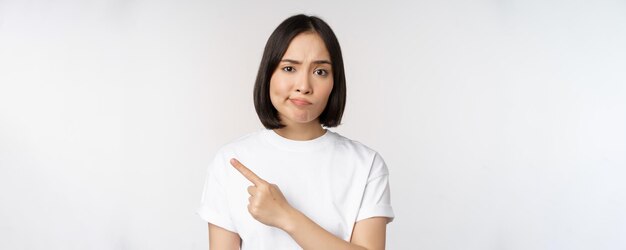 Chica asiática escéptica con camiseta blanca que señala el producto o el logotipo con una mueca decepcionada que no le gusta un