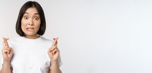 Chica asiática esperanzada cruza los dedos pidiendo deseo anticipando desear algo de pie en camiseta sobre blanco