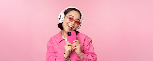 Chica asiática con estilo en auriculares escuchando música y tomando fotos en el teléfono móvil usando un teléfono inteligente de pie sobre fondo rosa