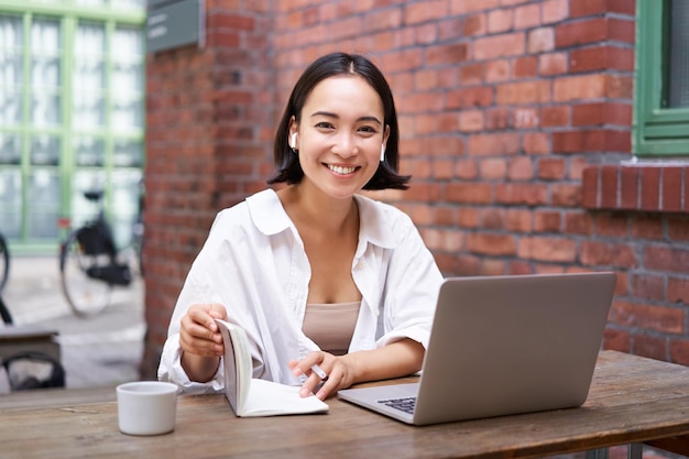 Chica asiática con estilo trabaja en coworking con una computadora portátil hace que las notas usen auriculares inalámbricos y las sonrisas se sientan