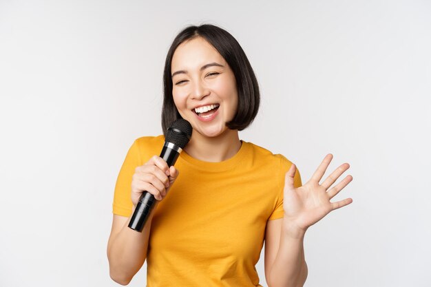 Chica asiática feliz bailando y cantando karaoke sosteniendo el micrófono en la mano divirtiéndose de pie sobre fondo blanco