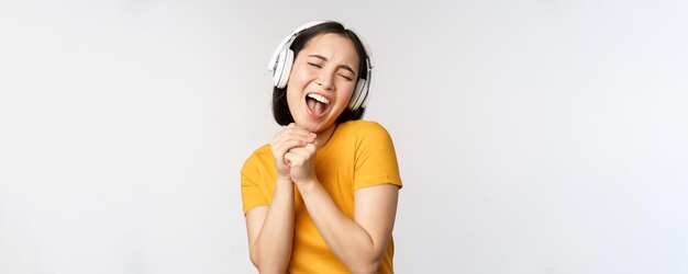 Chica asiática feliz bailando escuchando música en auriculares y sonriendo de pie en camiseta amarilla contra fondo blanco Copiar espacio