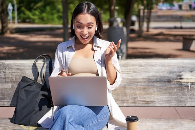 Chica asiática feliz chateando con una videollamada en una laptop sentada en un banco en el parque y comunicándose tal