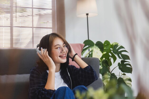 Chica asiática feliz con gafas escuchando música desde los auriculares en la sala de estar en casa Concepto de recreación en casa