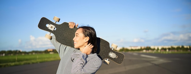 Foto gratuita chica asiática feliz y libre sosteniendo una tabla de crucero en los hombros y caminando hacia la cámara en vacío