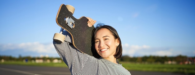Foto gratuita chica asiática feliz y libre sosteniendo una tabla de crucero en los hombros y caminando hacia la cámara en vacío
