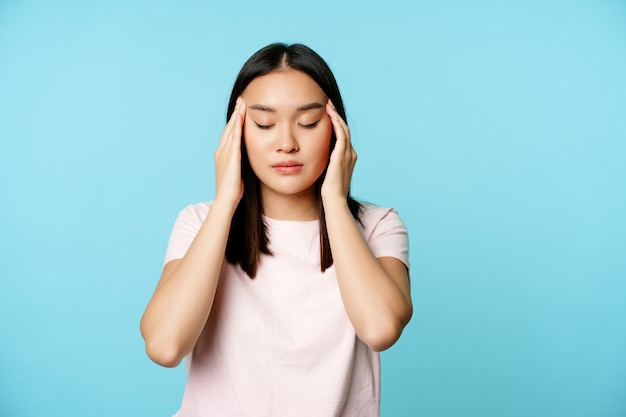 Chica asiática frotando, masajeando la cabeza con los dedos y los ojos cerrados, masaje de dolor de cabeza, con migraña, de pie sobre fondo azul.