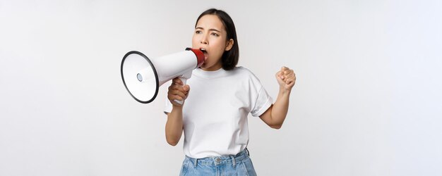 Chica asiática gritando al megáfono joven activista protestando usando un altavoz alto de pie sobre fondo blanco Copiar espacio