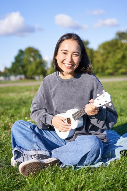 Chica asiática hipster positiva tocando el ukelele y haciendo muecas cantando y sintiéndose feliz