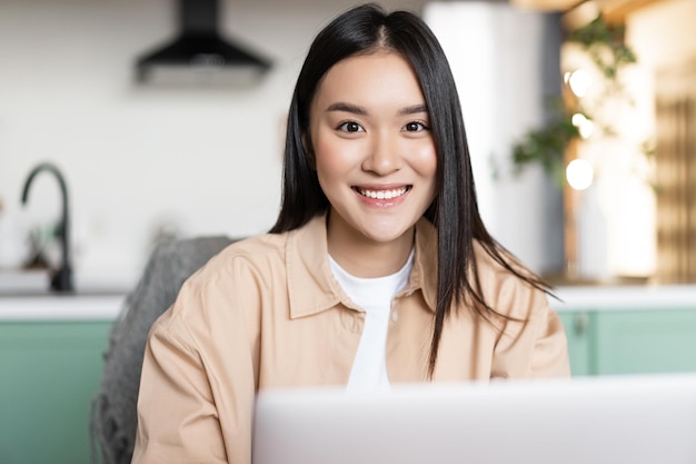 Chica asiática independiente sentada en casa y trabajando con la computadora portátil sonriendo a la cámara mujer joven que usa com ...