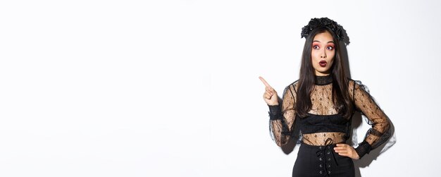Chica asiática intrigada celebrando halloween con vestido gótico negro y corona mirando y señalando la aleta