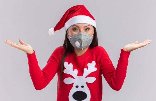 Chica asiática joven sorprendida con sombrero de Navidad con suéter y máscara médica extendiendo las manos aisladas sobre fondo blanco.
