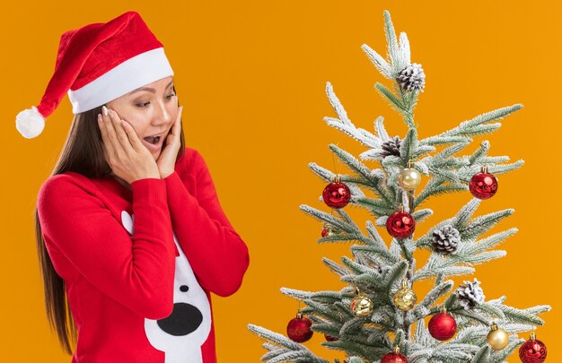 Chica asiática joven sorprendida con sombrero de navidad con suéter de pie cerca de árbol de navidad cubierto mejillas con manos aisladas sobre fondo naranja