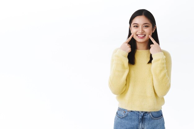 Chica asiática kawaii con cabello corto y oscuro, con un suéter amarillo, apuntando a los hoyuelos o mejillas y sonriendo, mostrando una piel perfecta sin imperfecciones, promueve el maquillaje coreano
