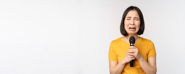 Chica asiática llorando cantando con el corazón roto en el micrófono sosteniendo el micrófono y haciendo muecas molesto de pie sobre lo que...