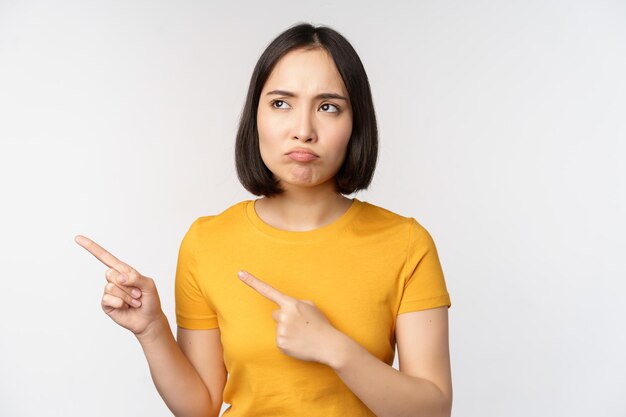 Una chica asiática malhumorada y malhumorada haciendo pucheros y mirando algo injusto señalando con el dedo a la izquierda en la marca del logotipo de la pancarta de pie con una camiseta amarilla sobre fondo blanco