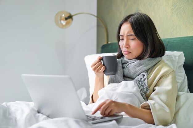 Foto gratuita chica asiática mirando la computadora portátil sintiéndose enferma resfriándose bebiendo té caliente para el dolor de garganta frunciendo el ceño como