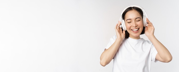 Chica asiática moderna bailando escuchando música con auriculares sonriendo feliz de pie en camiseta sobre fondo blanco.