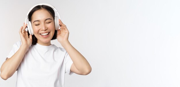 Chica asiática moderna bailando escuchando música con auriculares sonriendo feliz de pie en camiseta sobre Pentecostés