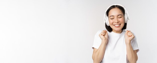 Chica asiática moderna bailando escuchando música con auriculares sonriendo feliz de pie en camiseta sobre Pentecostés