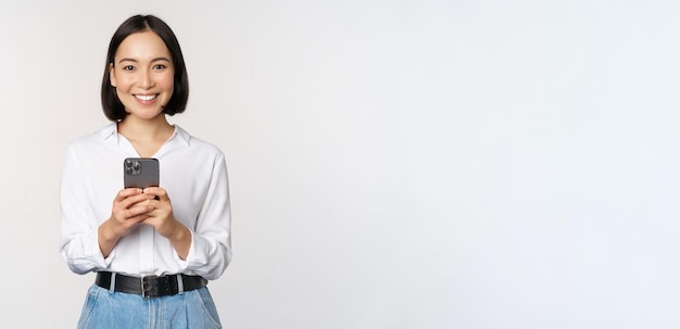 Chica asiática moderna y elegante usando una aplicación de teléfono móvil chateando por teléfono celular y sonriendo de pie en una blusa blanca contra el fondo del estudio