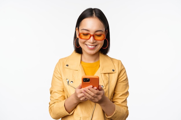 Chica asiática moderna con gafas de sol usando su teléfono móvil sonriendo y luciendo feliz posando sobre fondo blanco