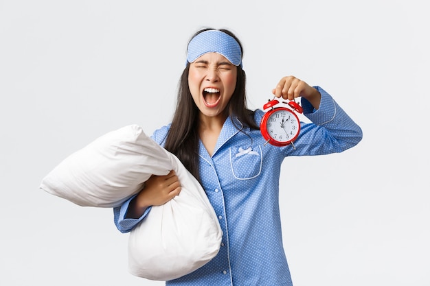 Chica asiática molesta y enojada en pijama azul y máscara para dormir gritando frustrada mientras se queda dormida, mostrando el despertador y gritando molesta, llegando tarde al trabajo, sosteniendo la almohada.