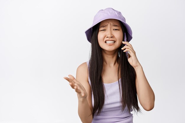 Chica asiática molesta que parece preocupada mientras recibe una llamada telefónica frunciendo el ceño y mirando angustiada con una conversación perturbadora de fondo blanco