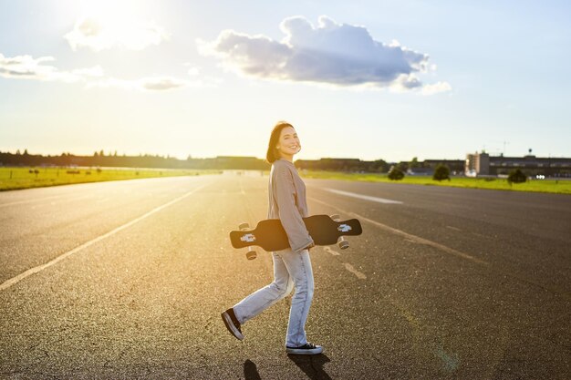 Chica asiática con monopatín de pie en la carretera durante la patinadora al atardecer posando con su crucero de tabla larga