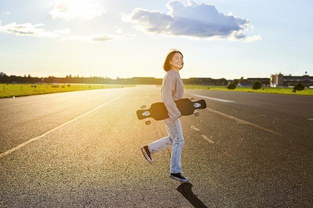 Foto gratuita chica asiática con monopatín de pie en la carretera durante la patinadora al atardecer posando con su crucero de tabla larga