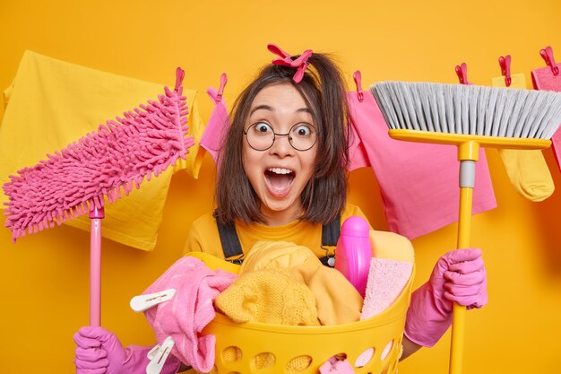 Chica asiática morena emocional sorprendida peina el cabello con pinzas para la ropa sostiene la fregona y el cepillo exclama en voz alta usa gafas redondas guantes de goma ocupada lavando poses contra el tendedero interior