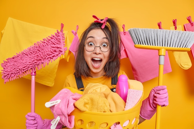 Chica asiática morena emocional sorprendida peina el cabello con pinzas para la ropa sostiene la fregona y el cepillo exclama en voz alta usa gafas redondas guantes de goma ocupada lavando poses contra el tendedero interior