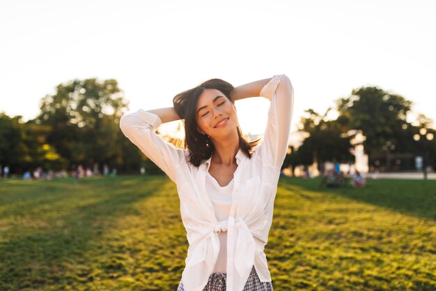 Chica asiática muy sonriente con camisa blanca cerrando los ojos soñadoramente mientras pasa felizmente el tiempo en el parque