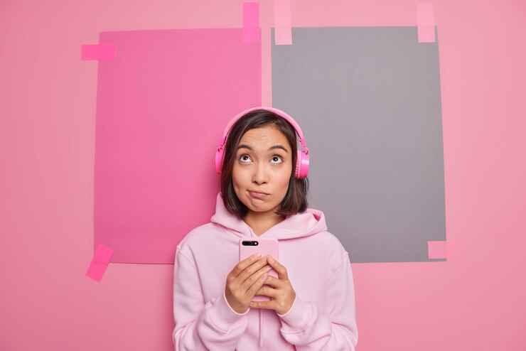 Foto gratuito a la chica asiática pensativa le gusta la nueva aplicación en el teléfono inteligente para las canciones de la lista de reproducción.