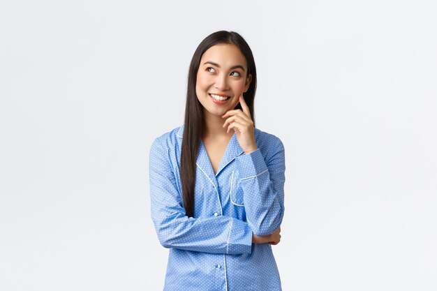 Chica asiática pensativa soñadora en pijama azul que tiene una idea interesante, mirando la esquina superior izquierda en la burbuja de comentarios, sonriendo complacida como pensando, soñando despierto antes de irse a la cama, fondo blanco.