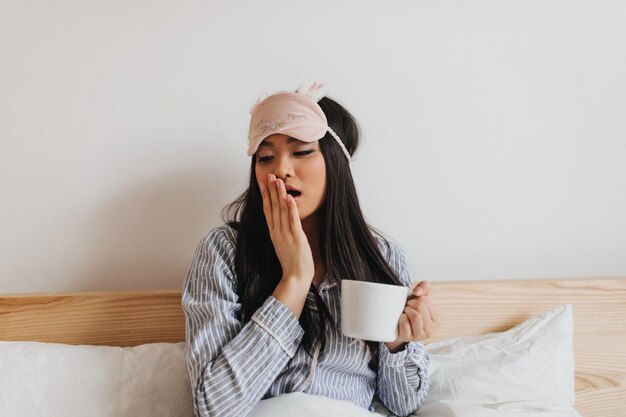 Chica asiática en pijama azul y lindo antifaz para dormir bosteza y posa con taza en la cama