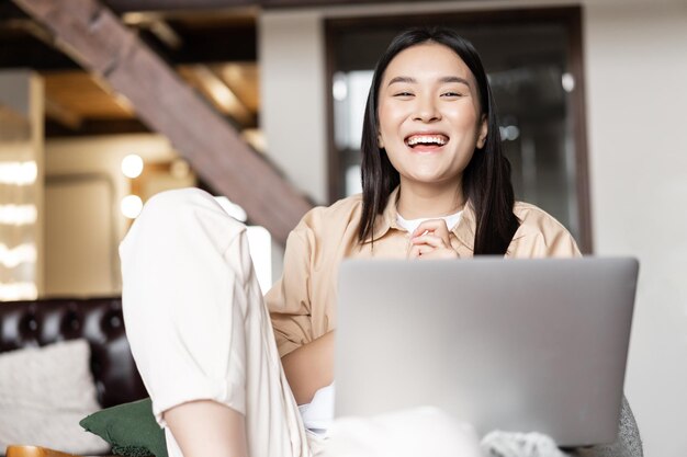 Chica asiática riendo relajándose en casa y viendo videos divertidos en la computadora portátil