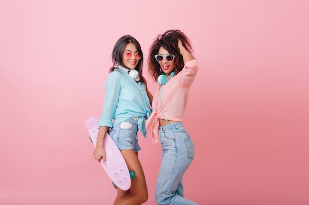 Chica asiática seria bronceada en gafas de sol rosas de pie con un amigo rizado africano y sosteniendo el patín. Elegante mujer mulata deportiva en jeans con auriculares jugando con el pelo