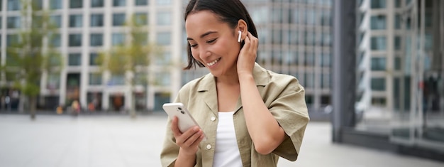 Foto gratuita una chica asiática sonriente escucha música con auriculares inalámbricos mira su teléfono eligiendo música o podcast