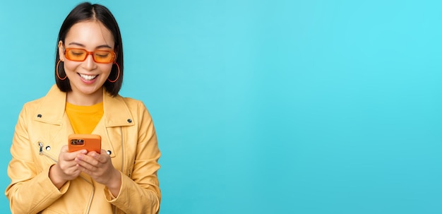 Chica asiática sonriente con gafas de sol usando una aplicación de teléfono inteligente sosteniendo un teléfono móvil de pie sobre un fondo azul