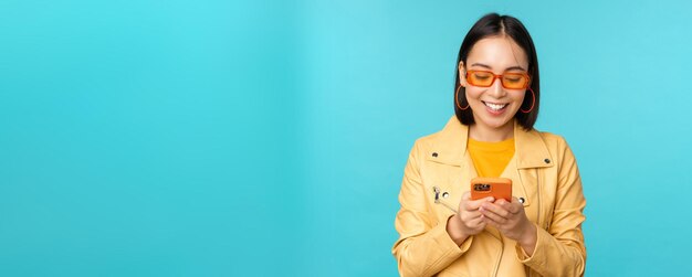 Chica asiática sonriente con gafas de sol usando una aplicación de teléfono inteligente sosteniendo un teléfono móvil de pie sobre un fondo azul