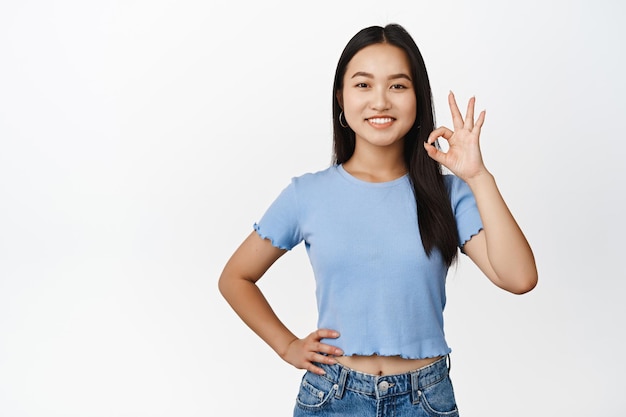 Una chica asiática sonriente muestra un gesto de estar bien para confirmar algo, decir que sí, haciendo un signo de Ok cero de pie sobre un fondo blanco